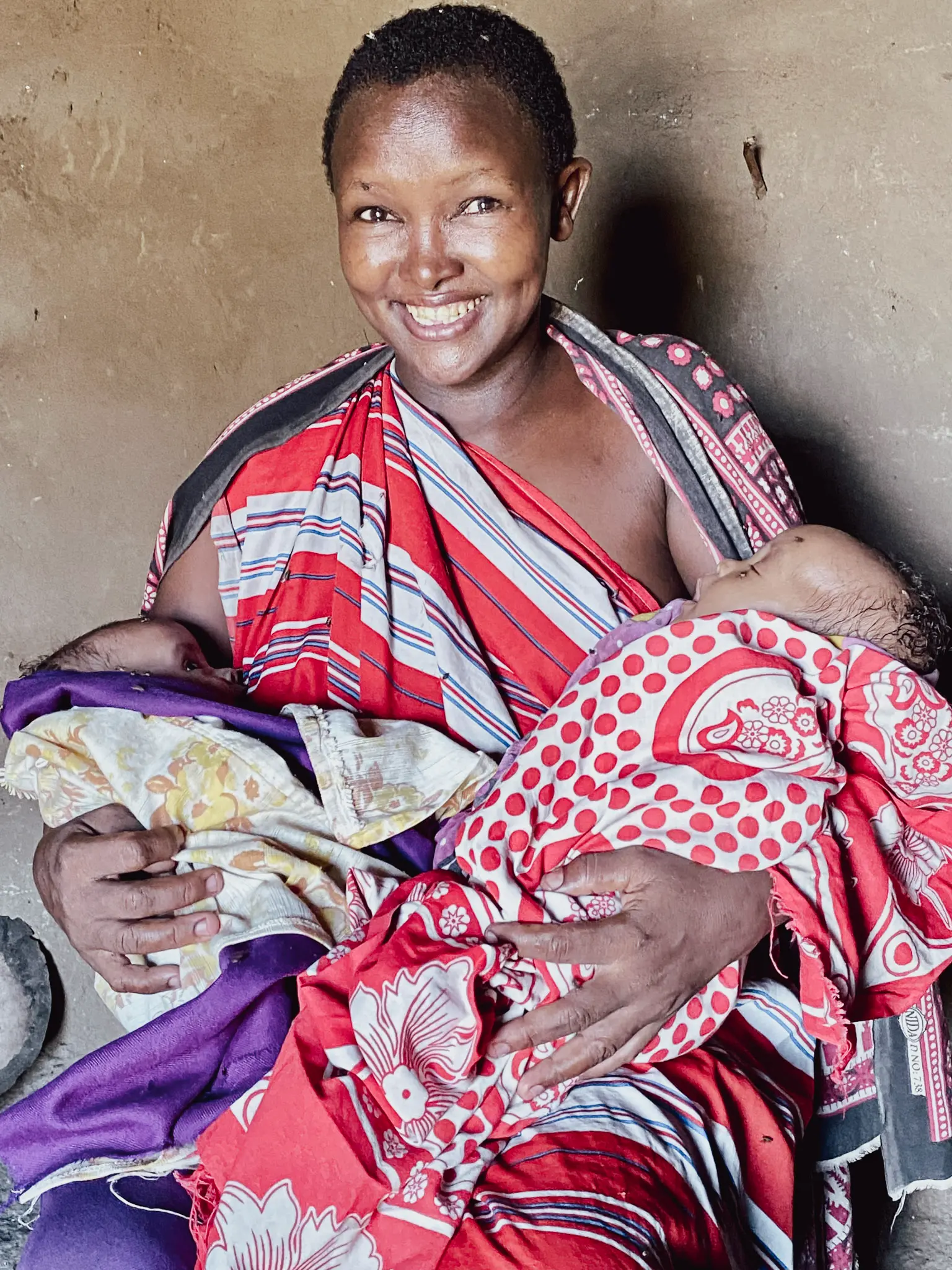 Woman holding two sleeping babies one per arm while wrapped in a red, white and gray piece of cloth, on baby wrapped in a purple and gold and the other in red and white patterned cloth.