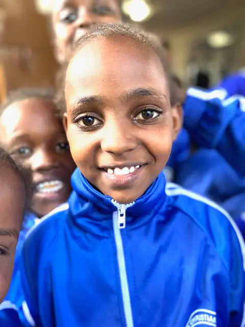 African Girl in her new blue track suit uniform with other kids trying to peek behind her.